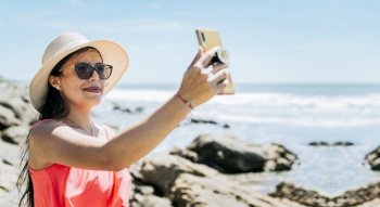 Girl in hat on the beach taking a selfie. Young woman on vacation taking photos on the beach, Smiling girl in hat taking photos on the beach. Beach vacation concept
