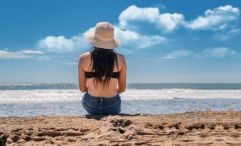 Girl in hat sitting with her back facing the sea, Young woman in hat sitting with her back looking at the sea, vacation girl concept, rear view of a girl looking at the sea