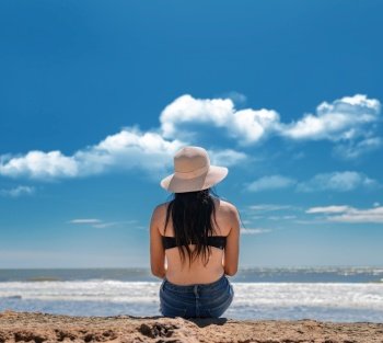 Young woman in hat sitting with her back looking at the sea, vacation girl concept, Girl in hat sitting with her back facing the sea, rear view of a girl looking at the sea