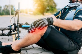 Concept of cyclist with knee injury outdoors. Cyclist with knee pain outdoors. Male cyclist sitting on the pavement with knee pain