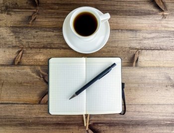 Coffee in a cup and a notepad with a pen, top view. Coffee in a cup with a saucer and a notebook with a pen on a wooden table.