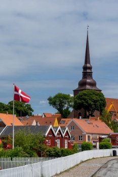 Nysted, Denmark - 11 June, 2021: red wooden houses and Danish flag with a tall church steeple behind