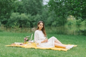 A beautiful young woman in a white dress drinks wine in the garden on a picnic. Summer vacation in solitude