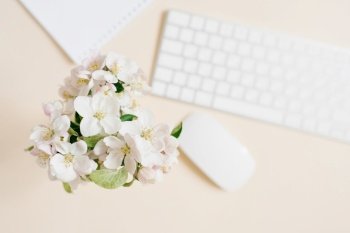 Keyboard, white sheet of paper and mouse and white apple flowers in a vase out of focus on a beige table. Layout and design concept. Flat lay.