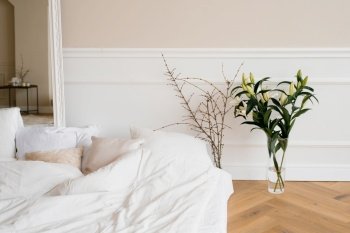A bright cozy bed with pillows and lily flowers in a vase, tree branches. Scandinavian interior of the house