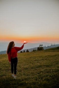 Pretty young woman standing on the mountain field at sunset and holding sun on the hand