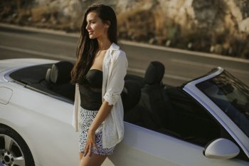Pretty young woman standing by the white cabriolet car on a sunny day by the sea