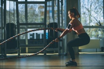 Pretty young woman with battle ropes exercise in the fitness gym