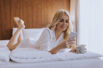 Happy womanl using a mobile phone while lying on the bed and drink morning coffee at home