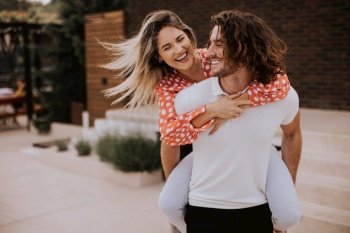 Handsome long hair man carrying the pretty young woman on his back in front of brick house