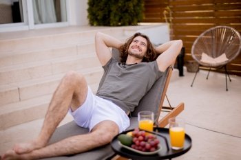 Handsome young man relaxing on deck chair with fresh fruits and cold orange juice by the swimming pool in the house backyard