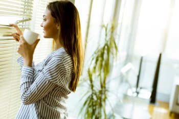 Attractive pretty young woman holding cup with hot tea or coffee and looking at the sunrise standing near the window in room