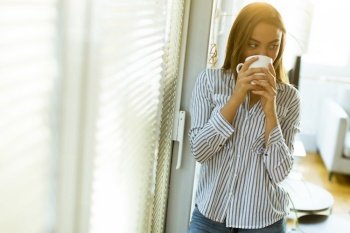 Attractive pretty young woman holding cup with hot tea or coffee and looking at the sunrise standing near the window in room