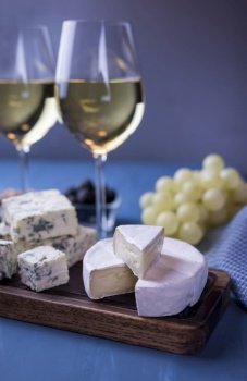 an appetizer assorted dor blue cheese and camembert on a wooden board with honey and grapes and with glasses of wine on a blue background