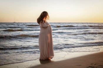 Young pregnant woman with a beautiful sea view on the background. Happy and calm pregnant woman with long hair and pink dress standig on the beach. Romantic view, ocean, sunset, maternity. Young pregnant woman with a beautiful sea view on the background. Happy and calm pregnant woman with long hair and pink dress standig on the beach. Romantic view, ocean, sunset, maternity.