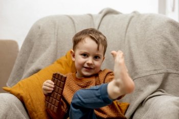 Little adorable boy sitting on the couch at home and eating chocolate bar. Child and sweets, sugar confectionery. Kid enjoy a delicious dessert. Preschool child with casual clothing. Positive emotion. Little adorable boy sitting on the couch at home and eating chocolate bar. Child and sweets, sugar confectionery. Kid enjoy a delicious dessert. Preschool child with casual clothing. Positive emotion.
