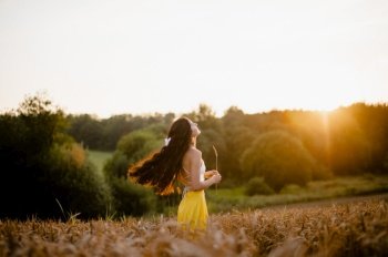 spring, sunset, wheat field, girl, evening, sun, yellow skirt, white t-shirt, white bow, hand with a spike, rustic style, girl’s smile, bread field, model, contour light. A girl in a yellow skirt stands in a field against the background of the setting sun