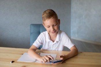 do homework, preparing for school, homework, look at the phone, peep the answer, the boy is sitting. white t-shirt, school period, open notebook, hold a pen, sit at the table. Cute boy writes in an open notebook and looks at the phone.