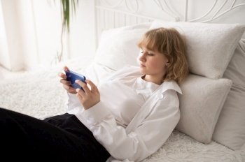 A happy European woman has a video call on her smartphone . The girl looks into the phone camera and smiles. White Scandinavian interior. Daylight through the window.. A cute girl in a white shirt and with a phone is lying on the bed. White interior