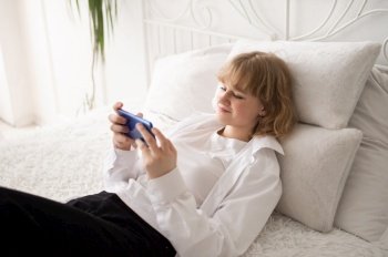 A happy European woman has a video call on her smartphone . The girl looks into the phone camera and smiles. White Scandinavian interior. Daylight through the window.. A cute girl in a white shirt and with a phone is lying on the bed. White interior