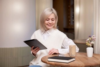 A cute girl in a shirt is sitting with a tablet and a diary in a cafe and drinking coffee. A cute girl is sitting with a tablet and a diary in a cafe and drinking coffee
