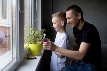 White T-shirt, dad hugs son, blonde, flower pot, watering flowers, house plants, window, window light. A cute boy is watering indoor plants. Dad next to his son near the window