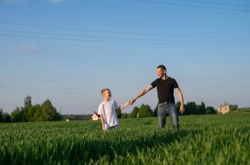 evening walk, white t-shirt, advertising space, children’s things, father and son, family weekend, stand in the field, run, jump, stand, nature, child’s smile, father, man, boy, child, adult. A cute child holds his father’s hand and they go into the field