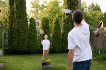 family games, house in the village, play tennis, badminton, teach the game, father and son, teach children, outdoor games, cooperation, advertising space, summer evening. Cheerful dad outside playing badminton with his son in a white T-shirt