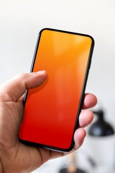 Hand holding a smartphone with blank red and orange screen. White office background.. Hand holding a smartphone with blank red and orange screen. Office background.
