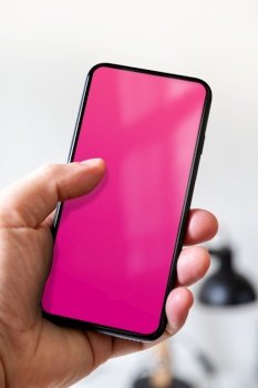 Hand holding a smartphone with blank pink screen. White office background.. Hand holding a smartphone with blank pink screen. Office background.