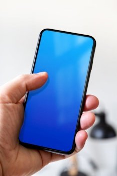 Hand holding a smartphone with blank blue screen. White office background.. Hand holding a smartphone with blank blue screen. Office background.