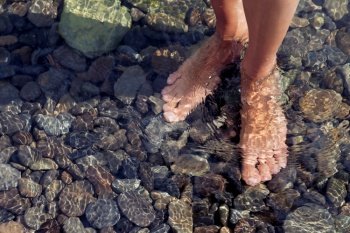 Woman’s legs ankle-deep in clear water. Ripple patterns reflect light on water over pebbles