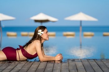 young woman in swimsuit relaxing on wooden edge of swimming pool with sea background