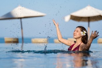 cheerful young woman in swimsuit playing water splashing in swimming pool with sea background