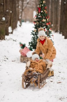 Children play with gifts next to a decorated Christmas tree in a winter snow-covered forest.. Children are sitting on gifts near the Christmas tree with balloons in the f