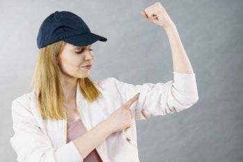 Young sporty woman wearing cap and sportswear showing her arm muscles enjoying workout results. Studio shot on grey background. Young woman showing her arm muscles