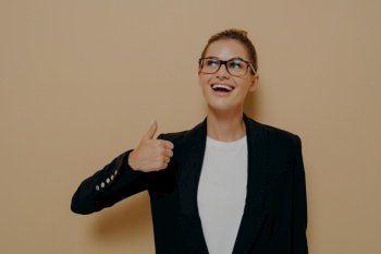 Portrait of satisfied female in eyeglasses smiling while showing thumb up with hand and looking upside with positive expression, demonstrating agreement while posing isolated over beige background. Portrait of satisfied female in eyeglasses smiling while showing thumb up with hand