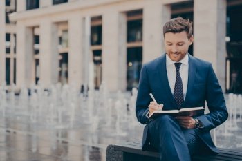 Formally dressed male office worker organising his day in notebook, writing it all down while sitting on bench, fountains working behind him, handsome businessman enjoying work outdoors on weekend. Formally dressed office worker organising his day in note book while working outdoors