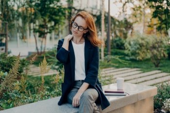Tired redhead woman takes rest in park suffers from neck pain sits on bench with takeaway coffee notebook digital device dressed formally poses outdoor. Businesswoman feels fatigue after busy day. Tired redhead woman takes rest in park suffers from neck pain sits on bench
