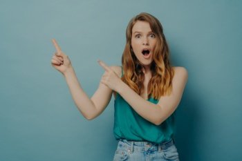 Surprised young female in casual wear pointing to right side with index fingers shocked by what she saw and looking at camera with mouth wide open isolated on blue studio background. Promotion concept. Surprised young woman in casual clothes pointing to right side with index fingers, isolated on blue