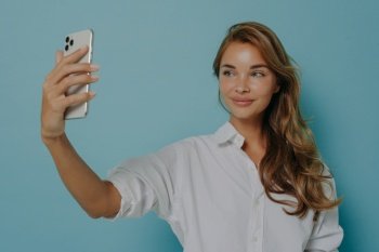 Indoor shot of good looking young woman with long hair takes selfie via modern smartphone takes photo of herself makes video call dressed in formal white shirt isolated over blue background.. Woman with long hair takes selfie via modern smartphone takes photo of herself makes video call