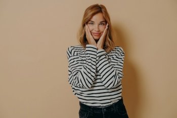 Cannot believe my eyes. Happy beautiful young woman in cozy striped longsleeve gently holding her face with palms, standing isolated over beige background and looking at camera with pleased smile. Happy beautiful young woman with pleased smile gently holding her face with palms