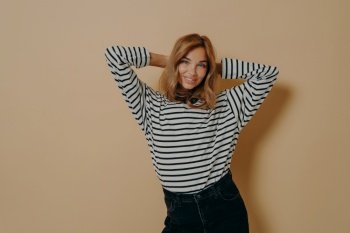 Half length shot of pretty satisfied young woman keeps hands behind head smiles gently wears striped jumper and black jeans isolated over beige background has natural beauty healthy clean skin. Half length shot of pretty satisfied young woman keeps hands behind head smiles gently poses indoor