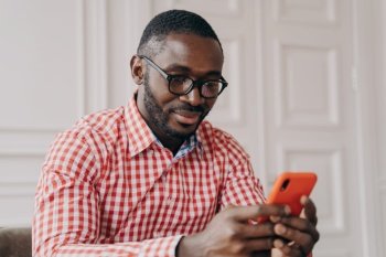 Cheerful millennial biracial man using smartphone, sitting at desk , young Afro american guy carefully looking on phone screen while typing urgent message, chatting online or searching in internet. Cheerful millennial biracial man using smartphone, sitting at desk