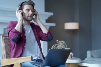 Young austrian guy sitting on wooden wicker chair with open laptop on his lap listening music in wireless headphones totally immersed in favorite, sounds closing eyes from pleasure of song melodies. Young german guy sitting on wicker chair with laptop on his lap listening music in headphones