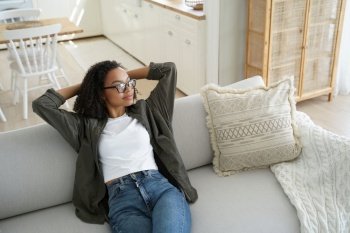 Calm african american young woman sitting relaxing on cozy sofa at home, holding hands behind head. Relaxed biracial girl dreaming, enjoying new house, resting on comfortable sofa in living room.. Calm african american young girl sitting relaxing dreaming on cozy couch in living room at home