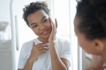 Mirror reflection of young attractive hispanic woman. Happy gorgeous african american girl applying cream to her face. Skin cleansing and treatment. Beauty routine and hygiene of tender teenage girl.. Happy african american girl is applying cream to her face. Skin cleansing and treatment.