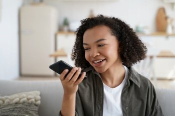 Friendly african american young girl sit on couch at home hold smartphone leave voice message. Modern biracial female chat with friends use loudspeaker virtual assistant search information on internet. Friendly african american young girl hold smartphone leave voice message or use virtual assistant