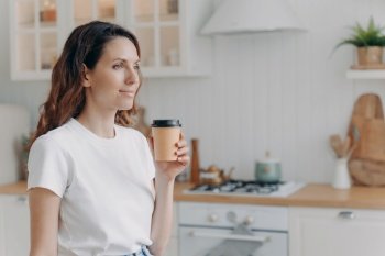 Relaxed smiling european woman with paper cup of coffee. Daily routine at home. Girl at kitchen in morning. Scandinavian stylish interior of apartment. White furniture, daylight.. Relaxed smiling woman with paper cup of coffee at home. Girl at kitchen in morning.