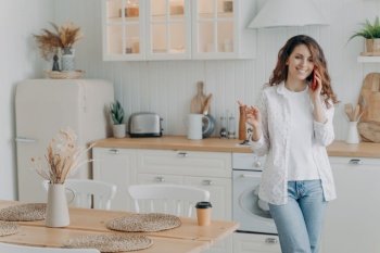 Young european woman talking on smartphone in her stylish kitchen at home. Happy attractive housewife has leisure. Modern white scandinavian interior. Stove, worktop and cuisine.. Happy housewife has leisure and talking on smartphone in kitchen. Modern scandinavian interior.